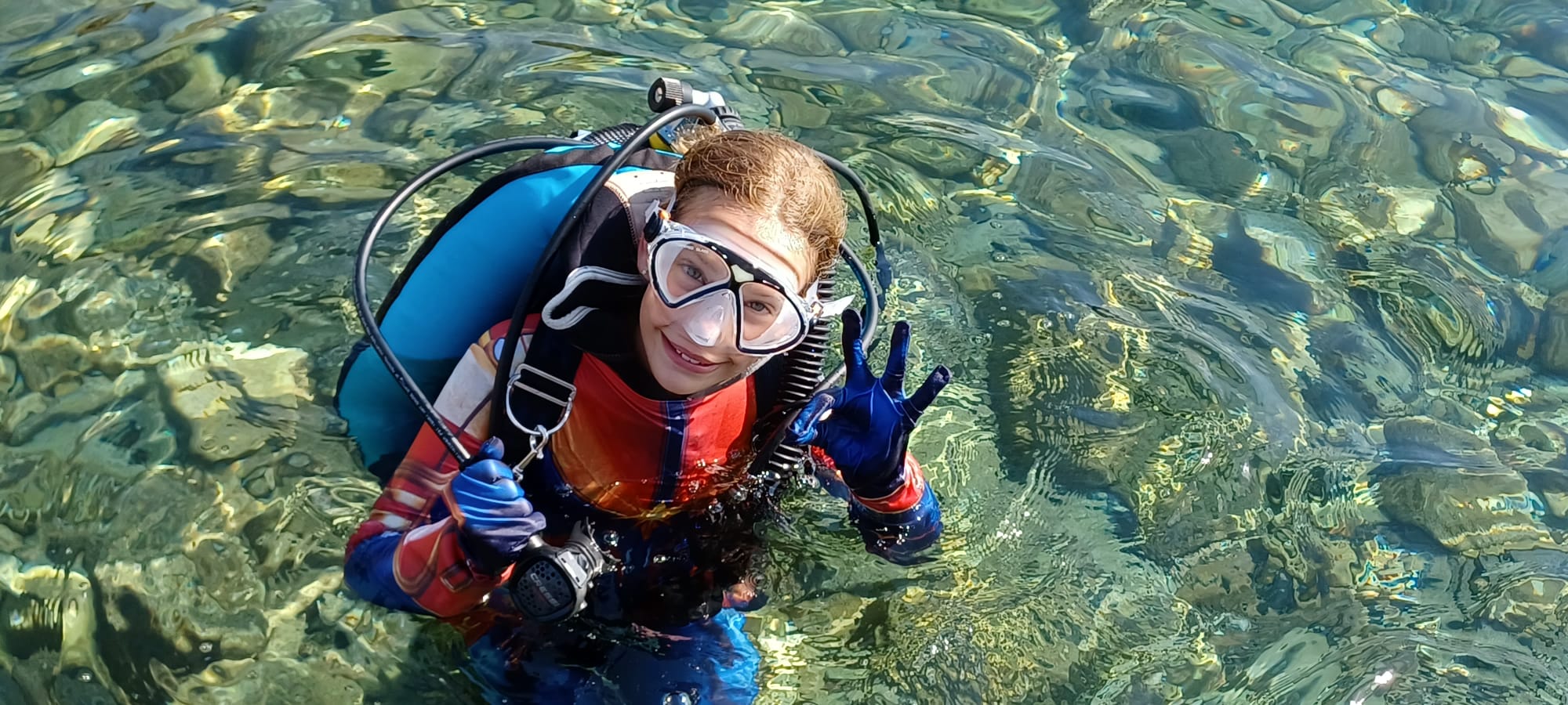 Nena amb equip de superheroi fent el senyal OK a l'aigua cristal·lina